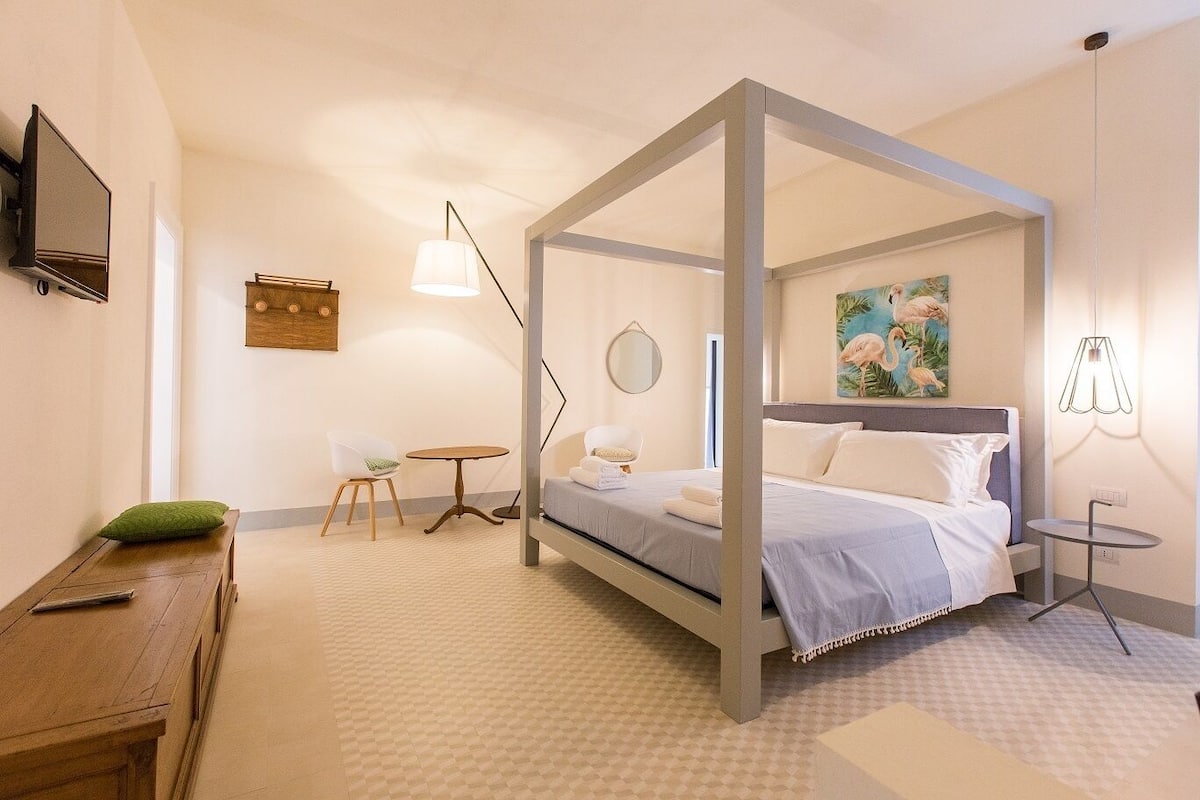 Una cama con dosel de estructura gris, un espejo redondo, una mesita pequeña y un cojín verde en un banco de madera.