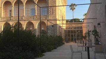 A courtyard with a stone building featuring arches and a tower. There are outdoor tables and chairs, and a glass door leading to an interior space.