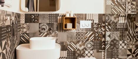 Une salle de bain avec des carreaux décorés, un lavabo blanc, un tabouret en bois, des toilettes blanches et deux lampes suspendues.