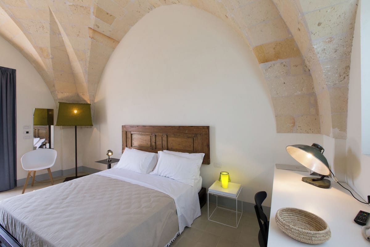 Un dormitorio con cabecera de madera, una cama, una mesita de noche con lámpara, una silla y una pequeña mesa con una cesta.