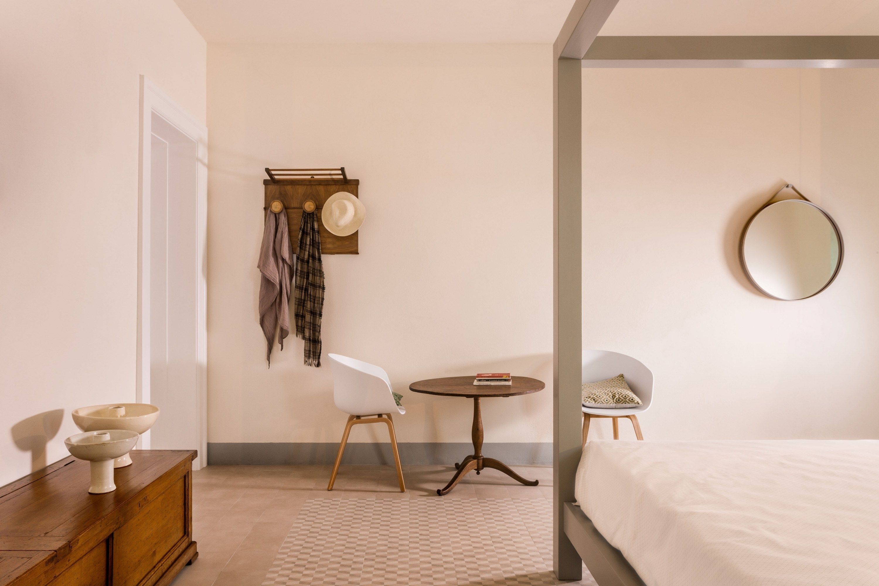 A bedroom with a bed, a round mirror, a small round table, two chairs, a wooden dresser, and a wall-mounted hat rack.
