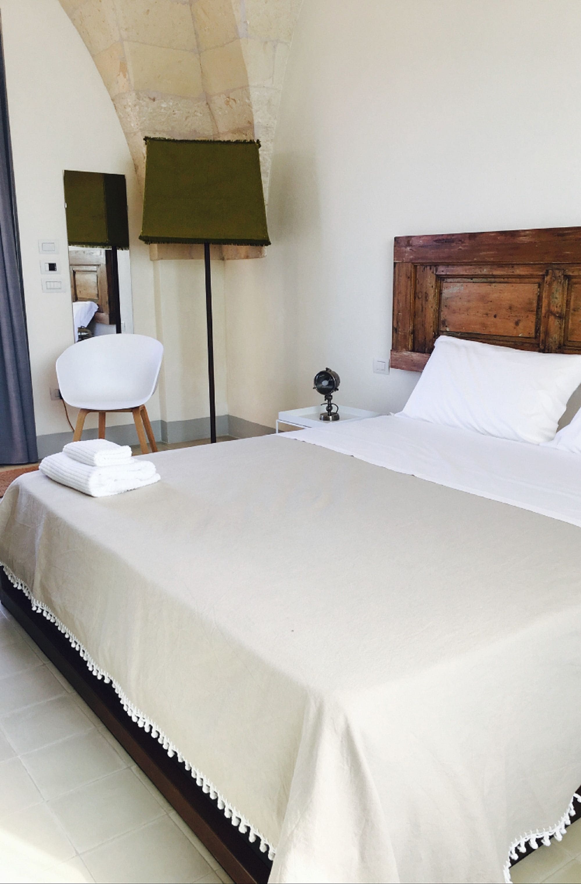 Una cama bien hecha con cubrecama blanco, cabecera de madera y mesita de noche con lámpara.