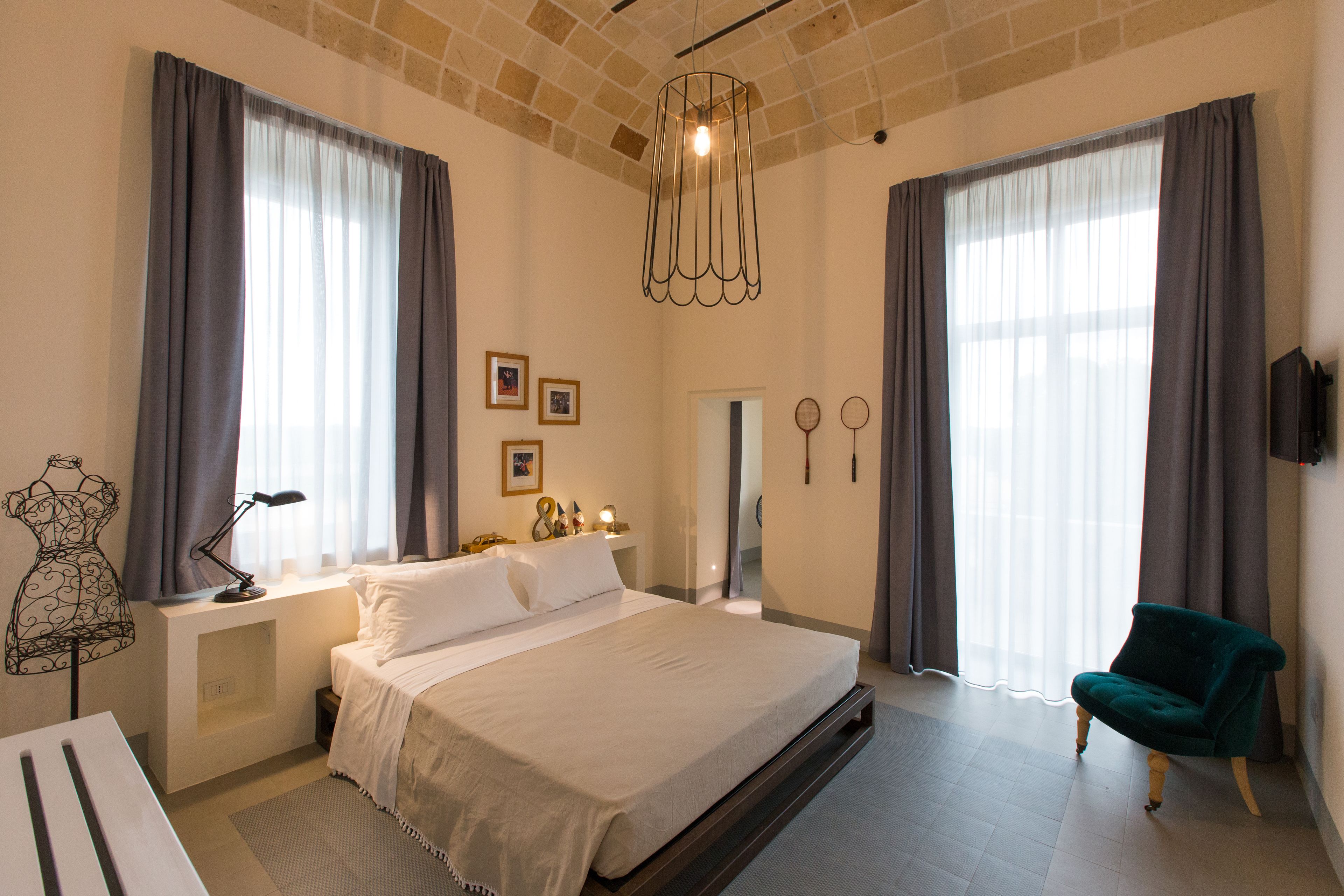 A spacious bedroom with a large bed, a desk, and a chair. There are two windows with curtains, a chandelier, and a wall-mounted TV.