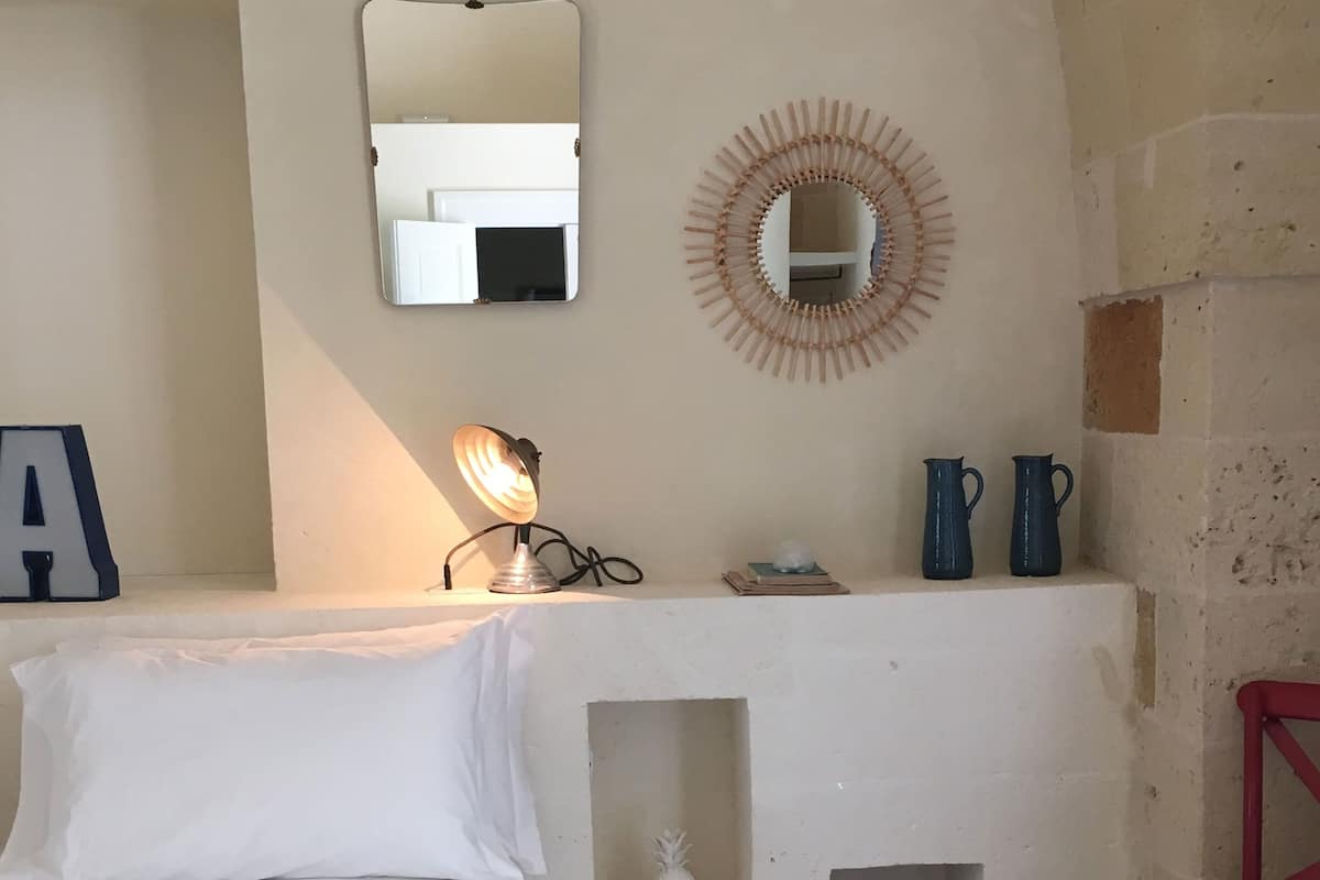 Un dormitorio con una cama, una mesita de noche con lámpara, un espejo y un pequeño estante con libros y una jarrón.