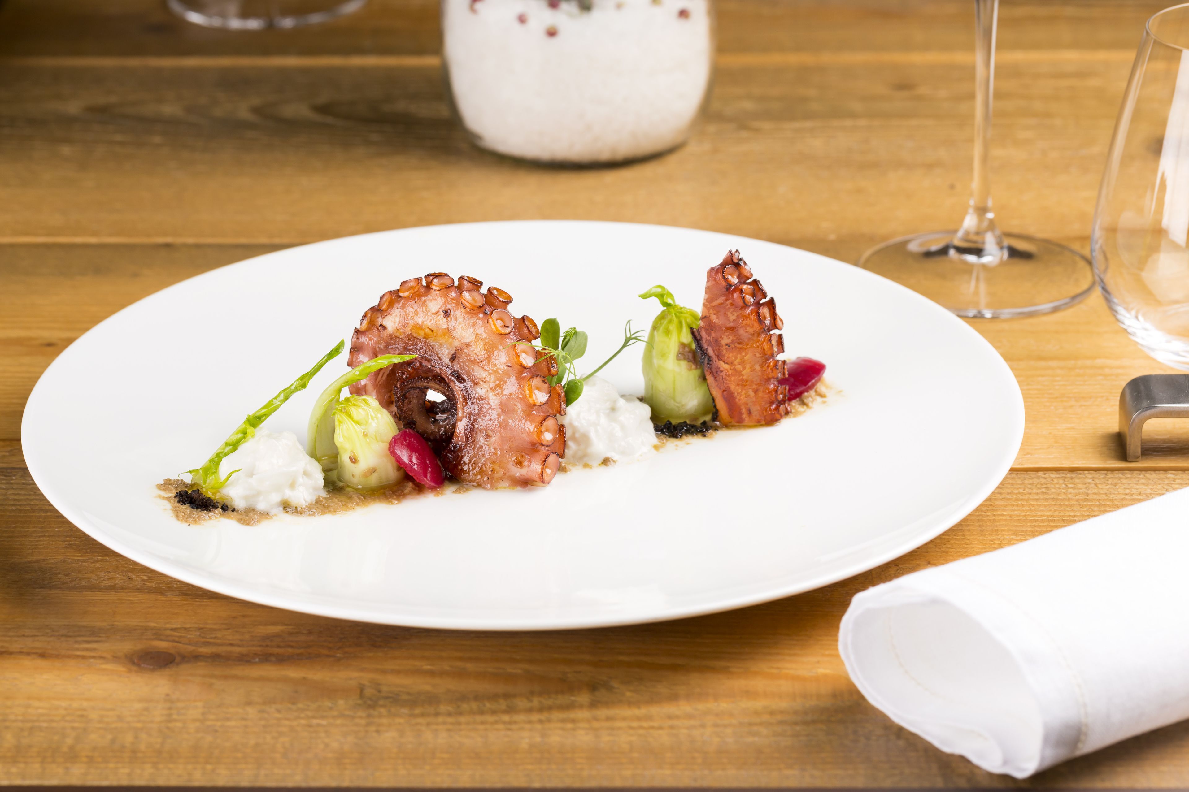 A white plate with octopus tentacles, greens, and a white sauce on a wooden table.