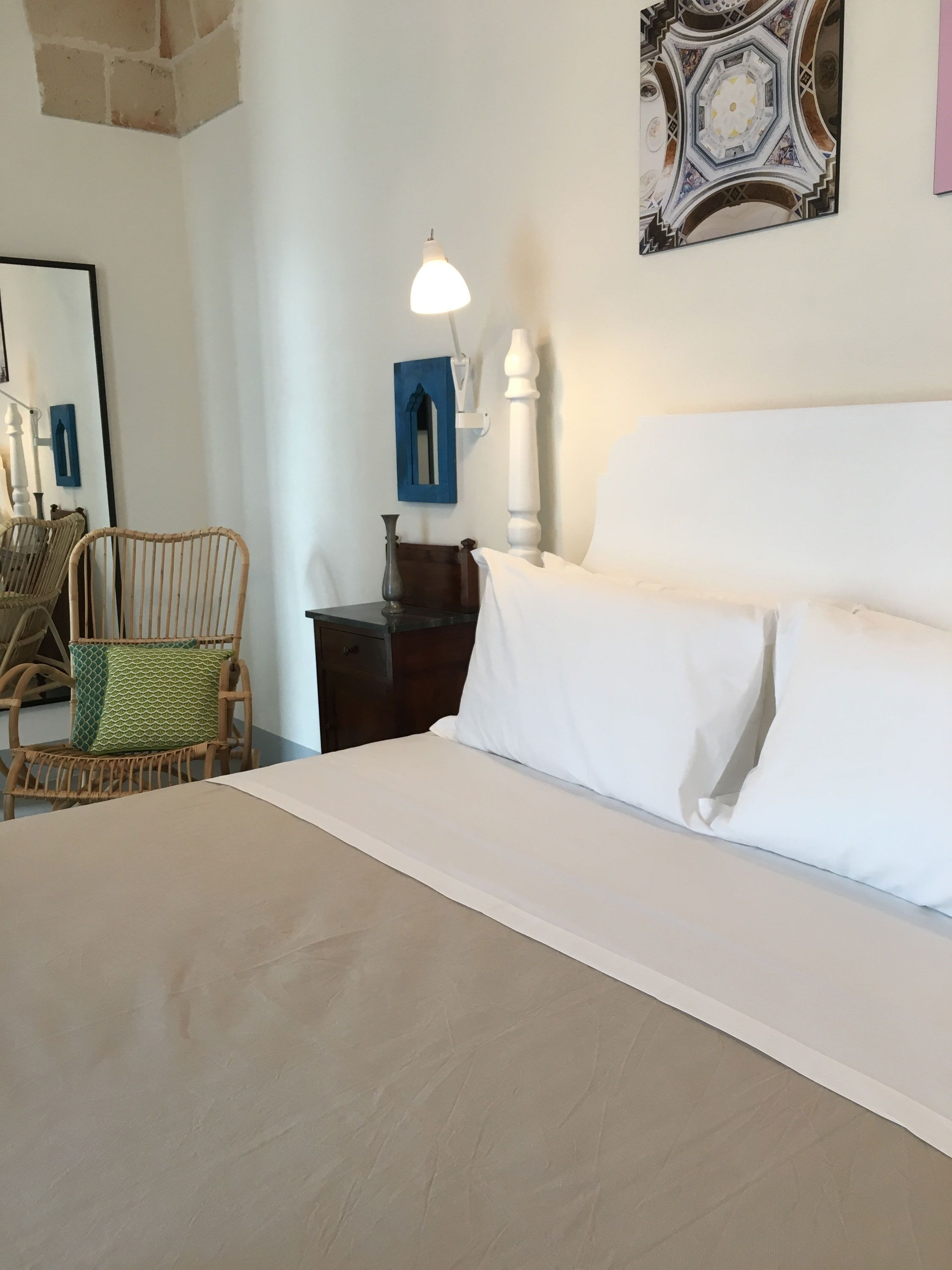 A bedroom with a large bed, a wicker chair, a nightstand, a lamp, and a wall-mounted mirror.