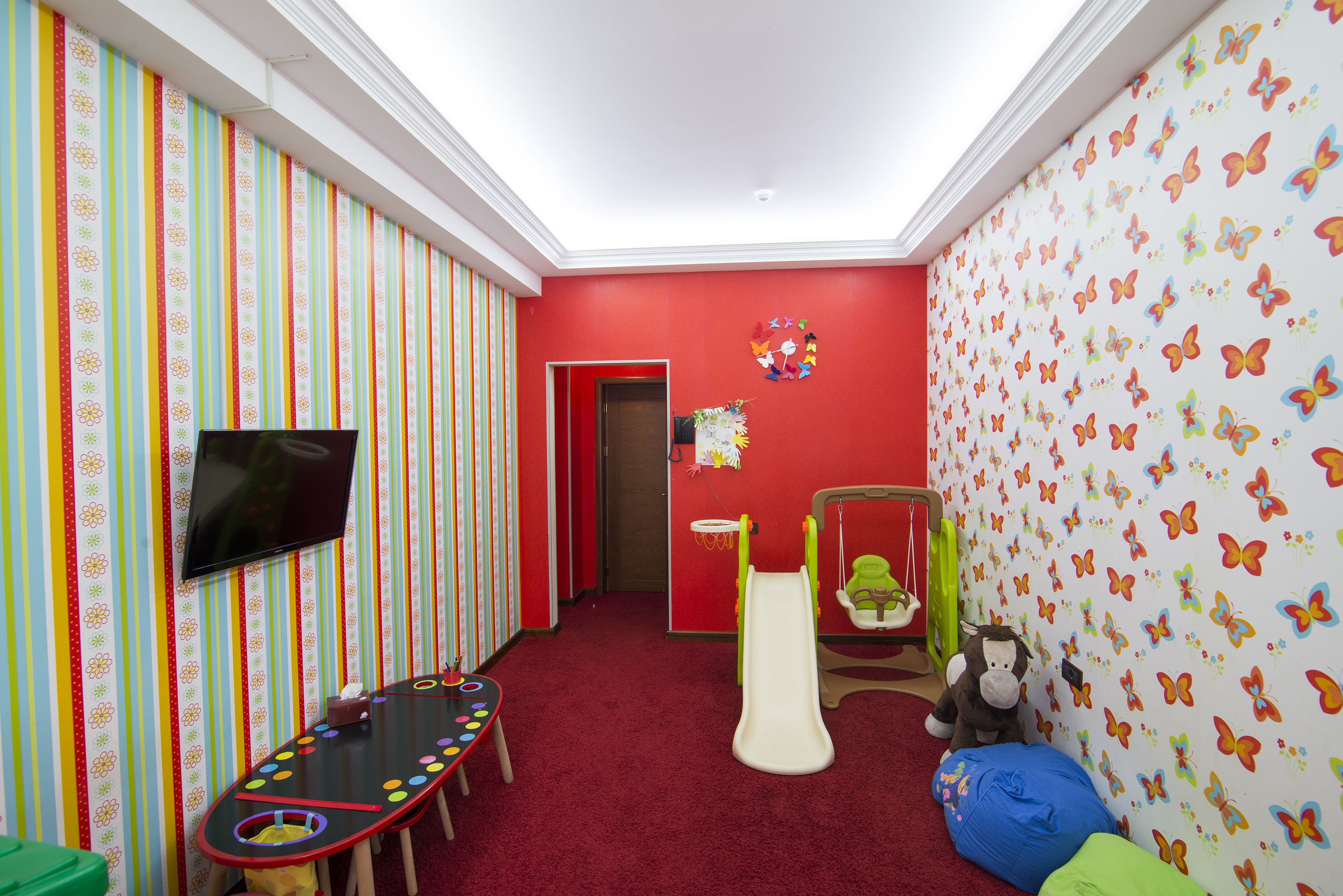 Una sala giochi con un muro rosso, uno scivolo, un tavolo dal motivo colorato e una televisione.