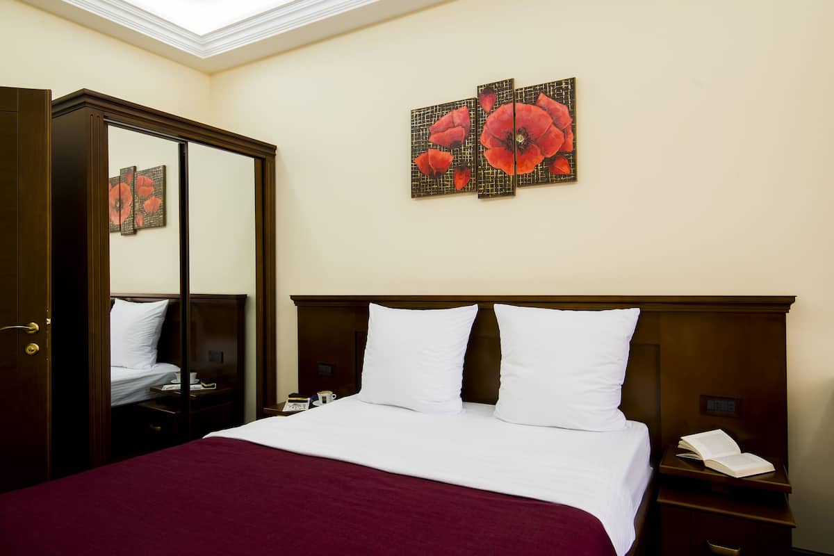A hotel room with a large bed, a wooden wardrobe, a bedside table with a lamp, and a wall-mounted artwork of red flowers.