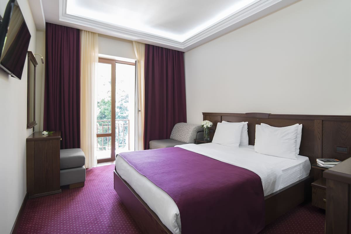 A hotel room with a bed, a nightstand, a TV, a desk, and a window with curtains.