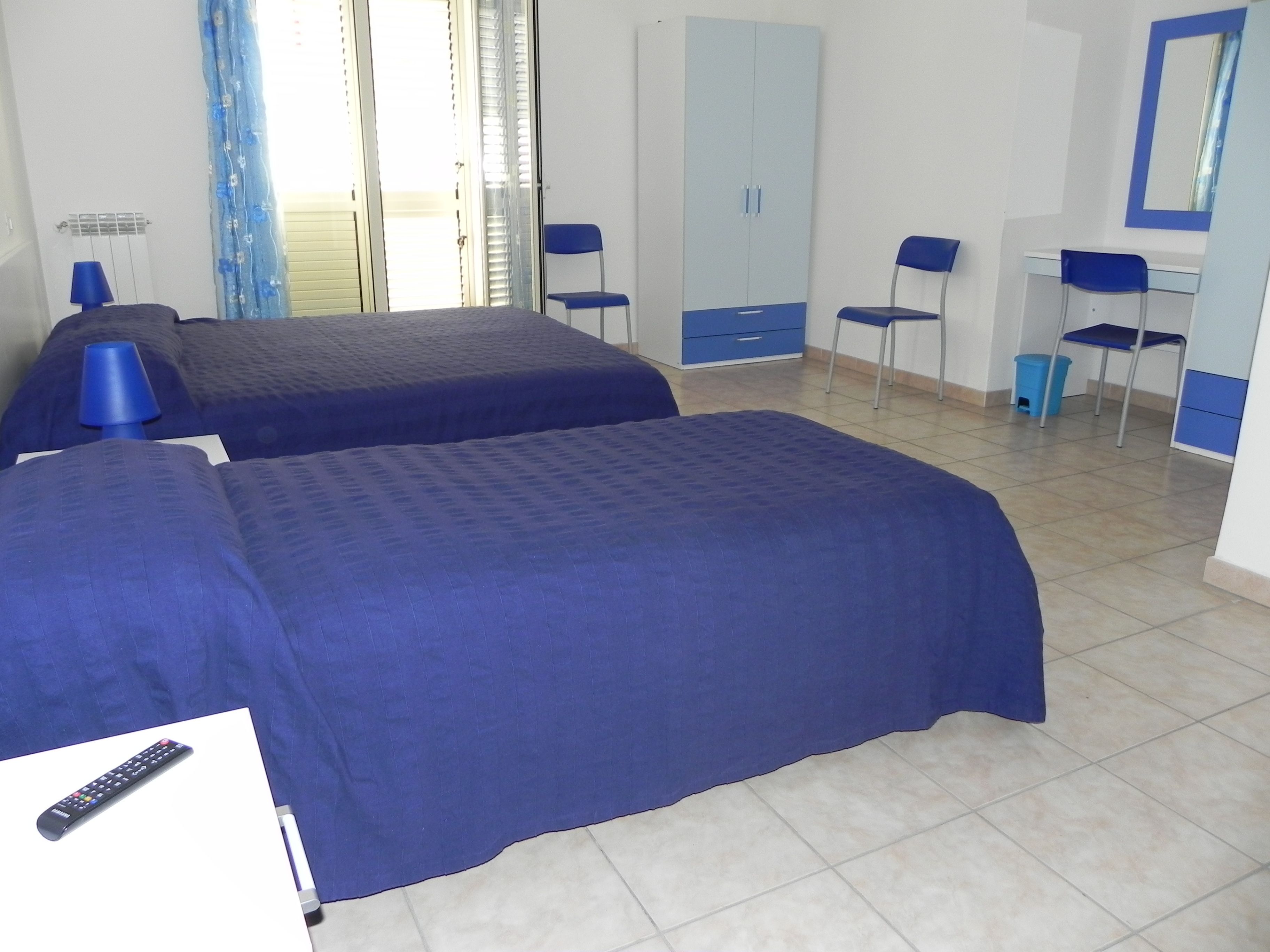 A hotel room with two beds, a wardrobe, a desk with chairs, and a window with curtains.