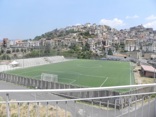 A soccer field with goalposts, surrounded by a metal fence. In the background, there is a densely packed hillside with various buildings. - Affittacamere Bellavista (Castiglione di Sicilia)