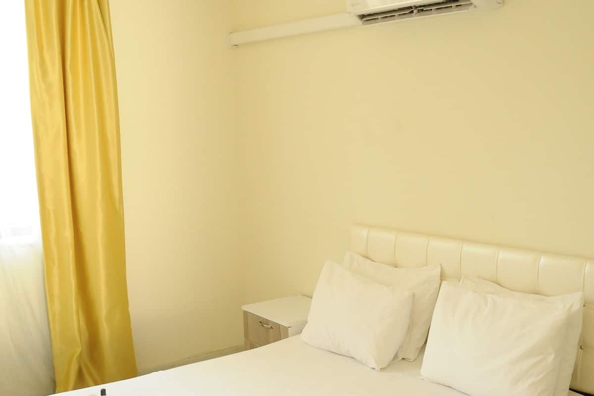 A hotel room with a bed, a nightstand, an air conditioning unit, and a yellow curtain.