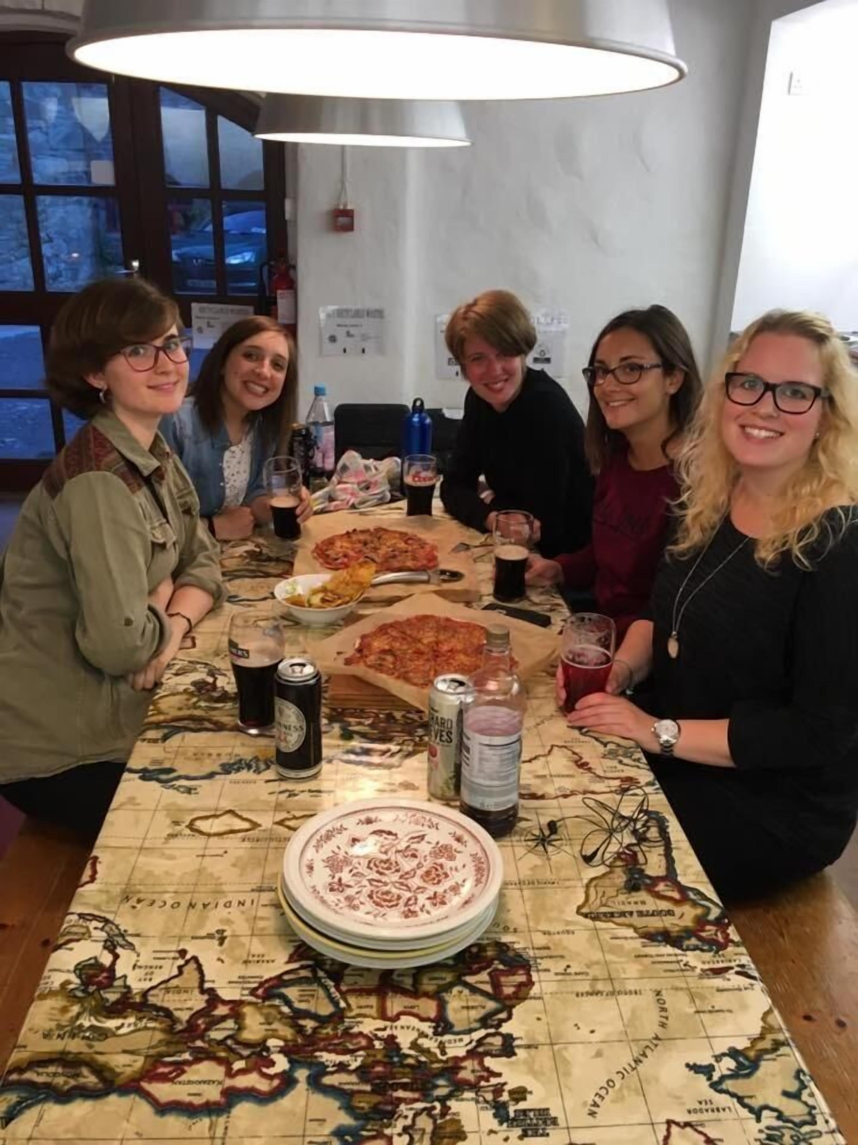 Um grupo de pessoas sentado à mesa, com pizza, bebidas e uma toalha de mesa com mapa.