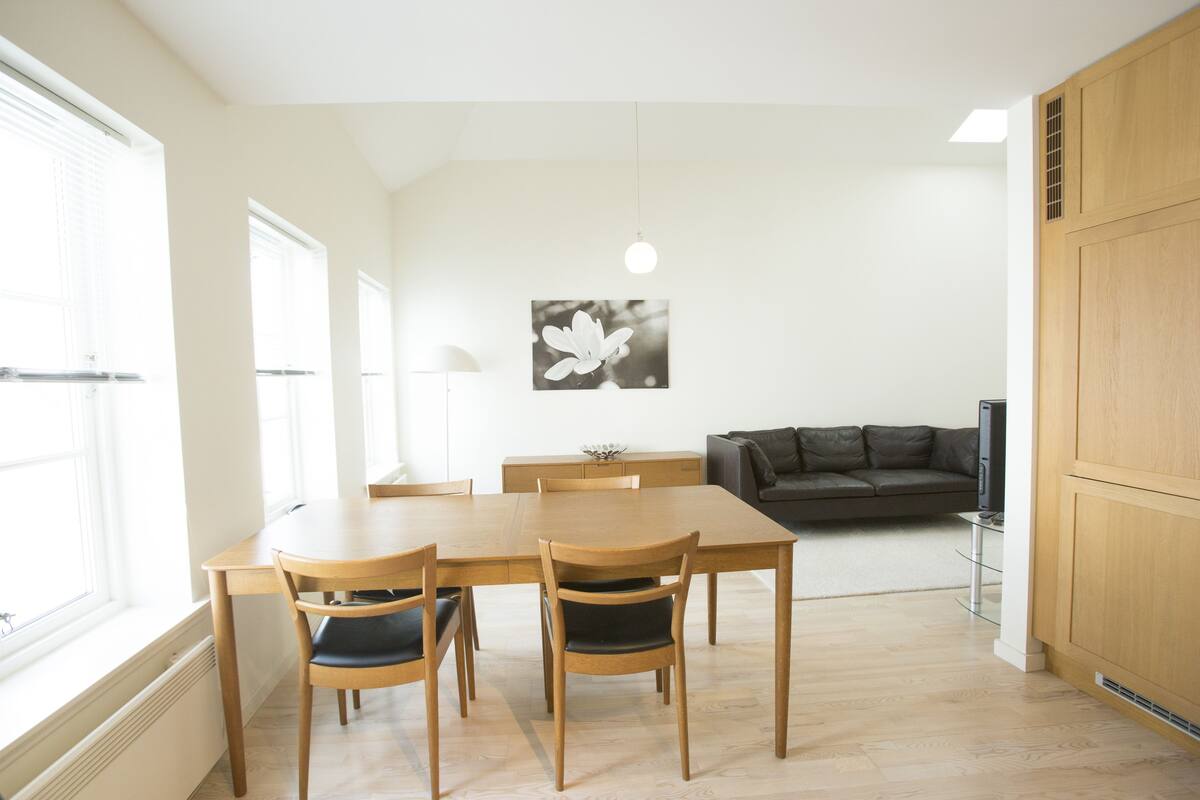 Ein Essbereich mit Holztisch und -stühlen, einem schwarz-weißen Blumenbild an der Wand und einer dunkelgrauen Couch im Hintergrund.