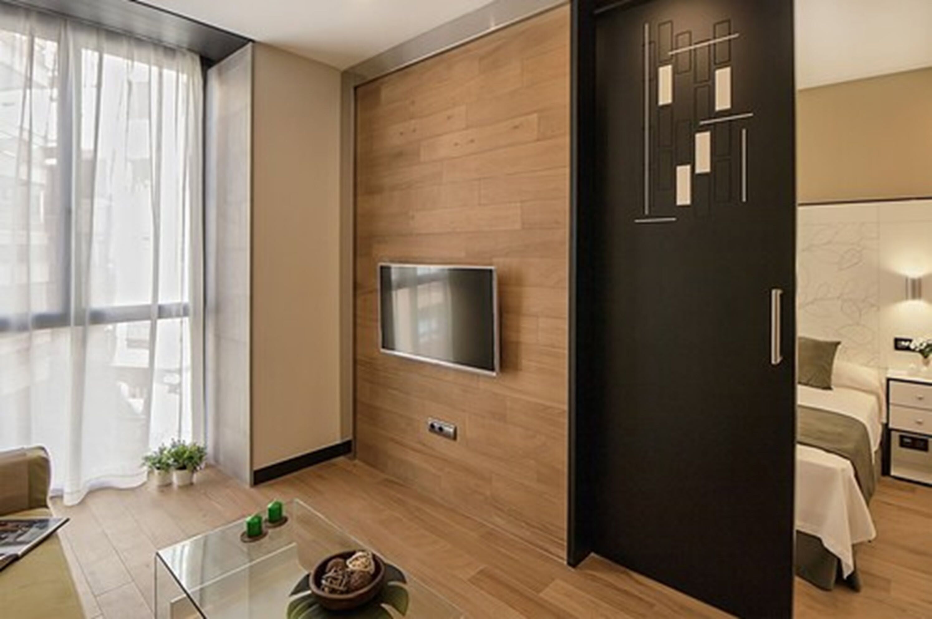 Une chambre d’hôtel moderne avec un plancher en bois, un téléviseur à écran plat fixé au mur, un lit avec une tête de lit blanche et une petite table de chevet avec une lampe.
