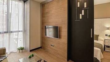 Une chambre d’hôtel moderne avec un plancher en bois, un téléviseur à écran plat fixé au mur, un lit avec une tête de lit blanche et une petite table de chevet avec une lampe.