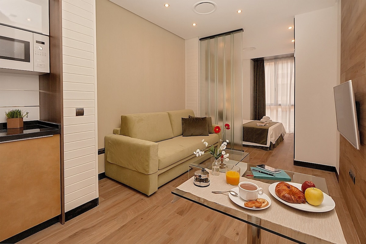 Ein modernes Hotelzimmer mit Küchenzeile, einer Couch, einem Essbereich mit Frühstück und einem Fernseher.