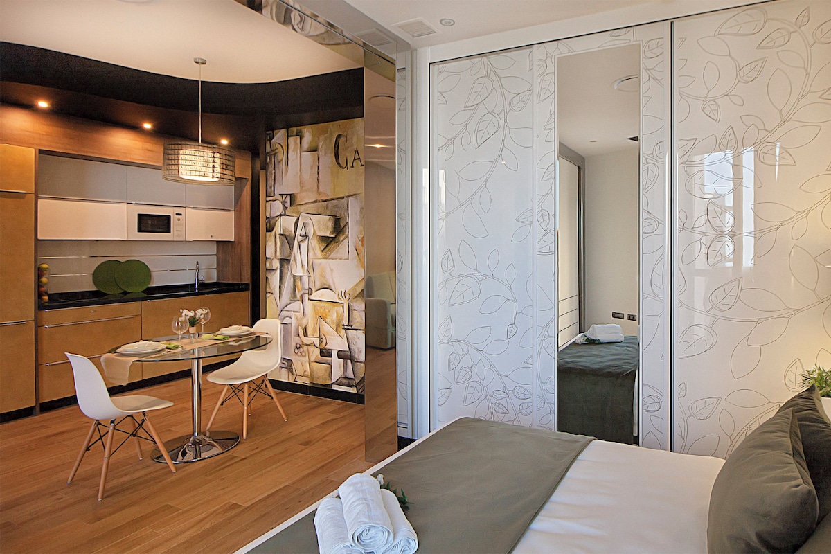 A modern hotel room with a bed, a seating area with a table and chairs, a kitchenette, and a bathroom visible through a sliding door.