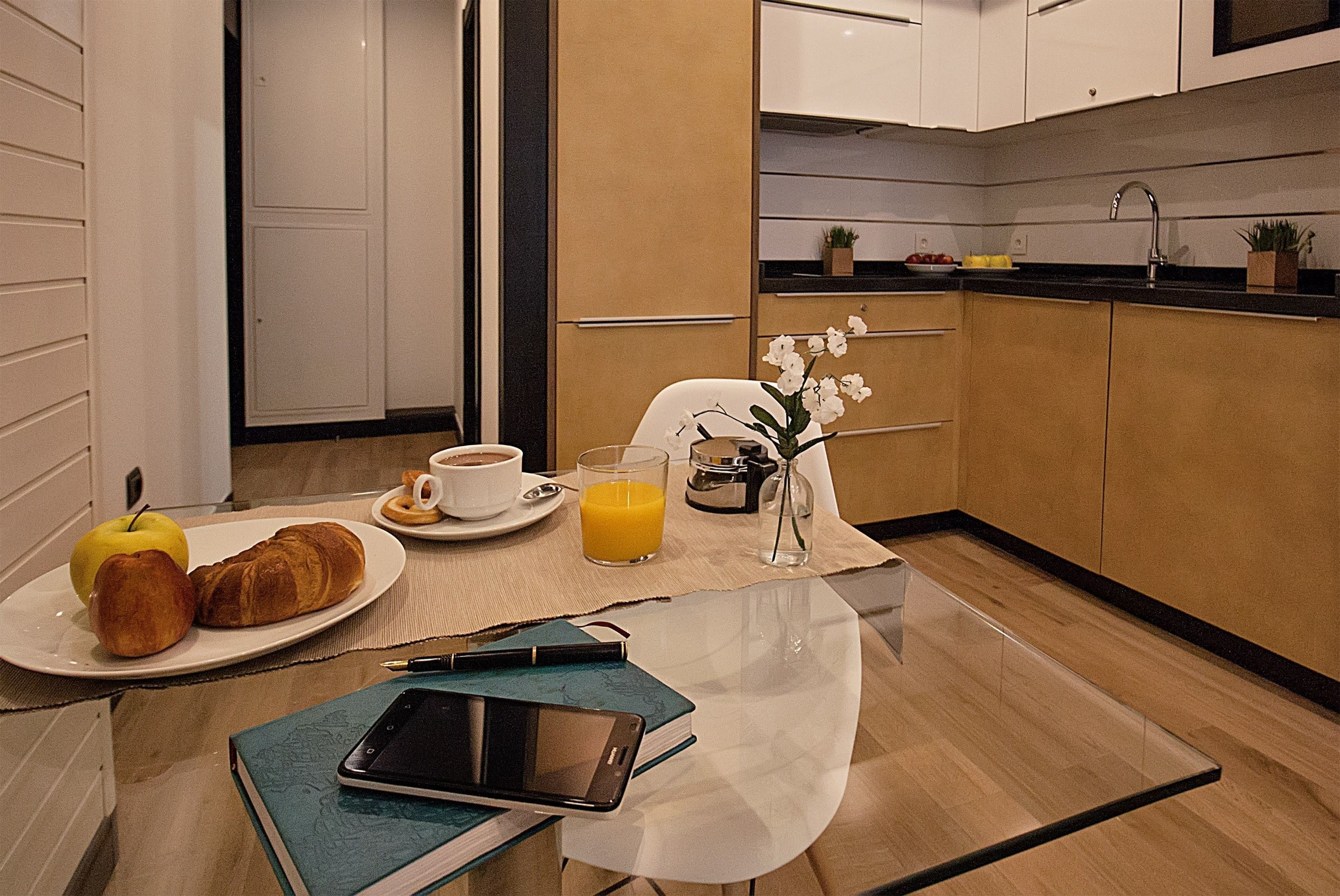 A breakfast setup with croissants, an apple, a glass of orange juice, a cup of coffee, and a tablet on a table.