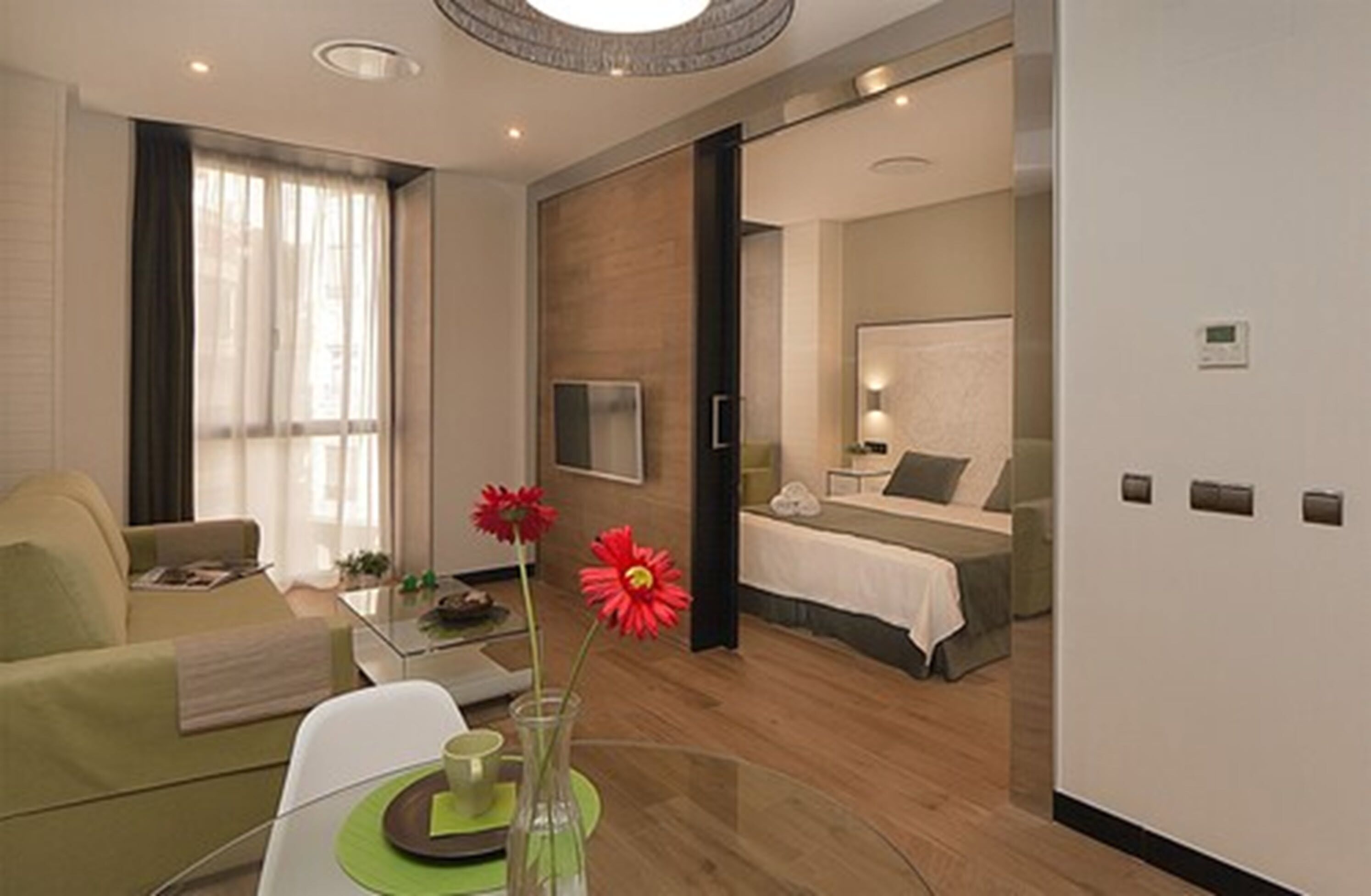 A modern hotel room with a sofa, a dining table with a vase of flowers, a bed with a white headboard, and a large mirror.
