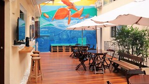 An outdoor dining area with wooden tables and chairs, a mural of a surfer, and umbrellas providing shade. - Hostel da Barra (Rio de Janeiro)