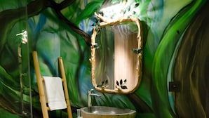 Une salle de bain décorée d’une fresque murale aux motifs verts, avec une chaise en bois et un grand miroir au-dessus d’un lavabo.