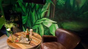 Une pièce avec une table basse ronde en bois, un fauteuil en cuir marron, un téléviseur fixé au mur et une fresque murale représentant des feuilles vertes.