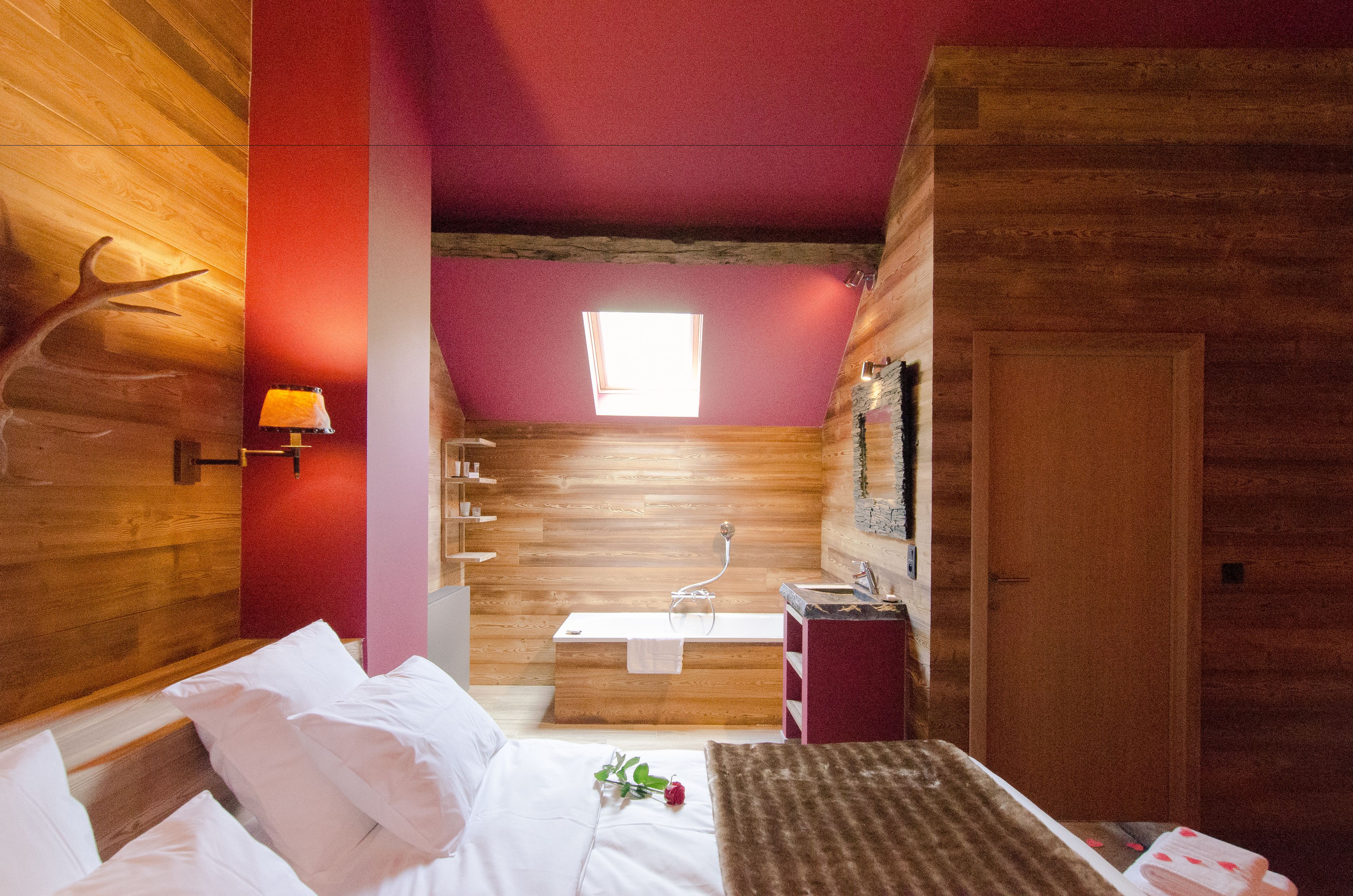 A wooden cabin bedroom with a bed, a small window, a wooden wall with a deer antler decoration, and a bathroom with a sink and mirror.