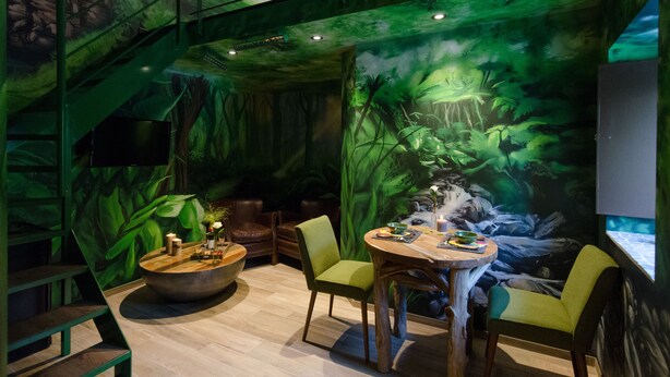 Un restaurant décoré d’une fresque murale sur le thème de la jungle, avec des meubles en bois et un escalier.