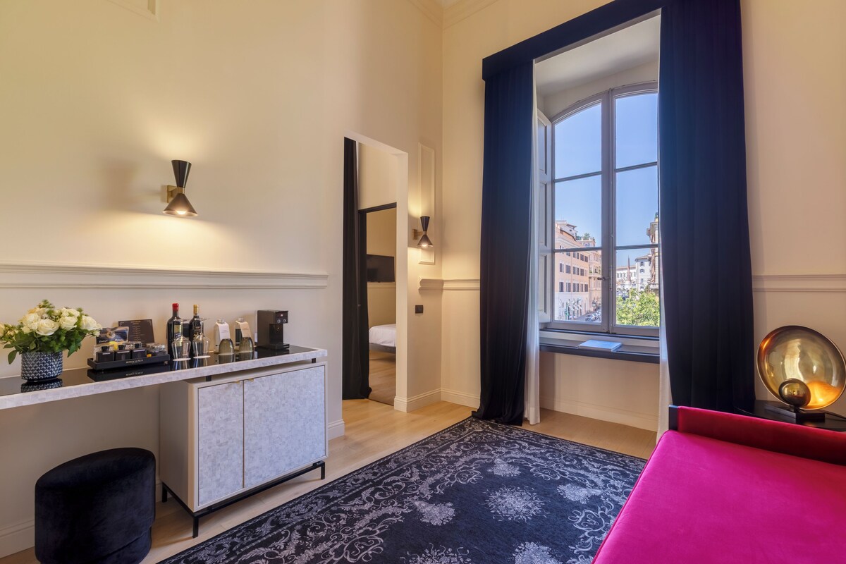 A hotel room with a bed, a desk with a coffee machine, a minibar, and a view of the city through a large window.