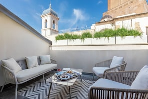 A rooftop terrace with a white sofa, two wicker chairs, a table with a tray of food, and a view of a church tower and building.