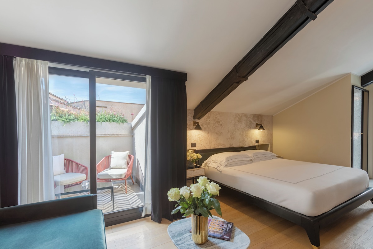 A hotel room with a bed, a small table with a vase of flowers, a chair, and a view of the outside.