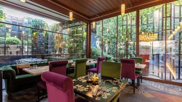 A restaurant with a view of lush greenery through large windows, wooden ceiling beams, and colorful upholstered chairs.