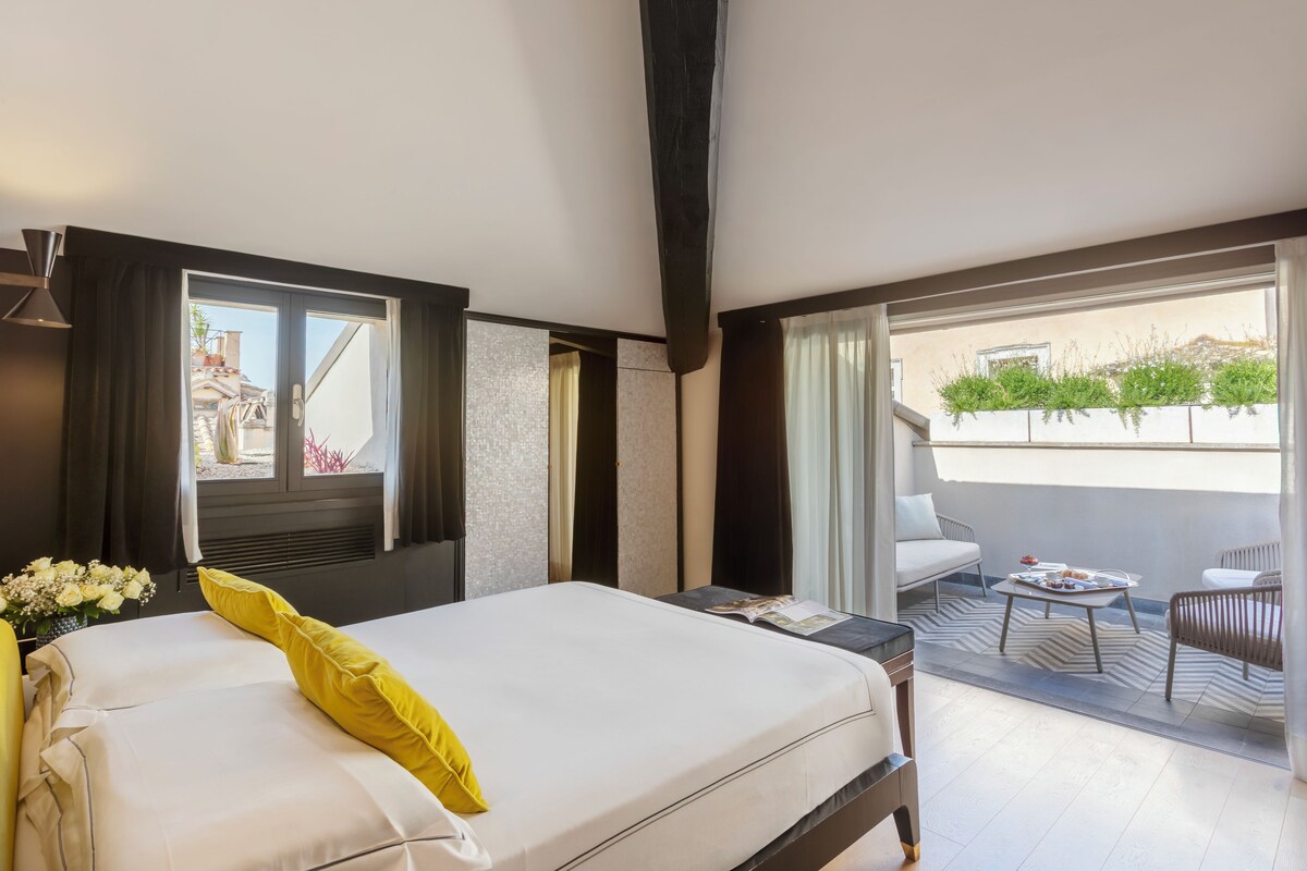 A hotel room with a bed, a small table with a magazine, a balcony with a chair, and a view of buildings and greenery.