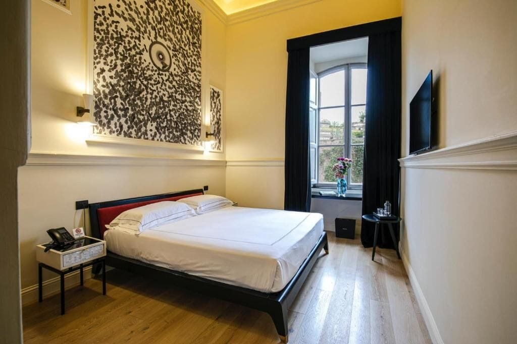 A hotel room with a bed, a nightstand with a phone, a wall art piece, a window with curtains, and a TV mounted on the wall.