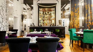 Un restaurante con zona de bar, mesas con sillas tapizadas en morado y verde, y una amplia exhibición de botellas al fondo.