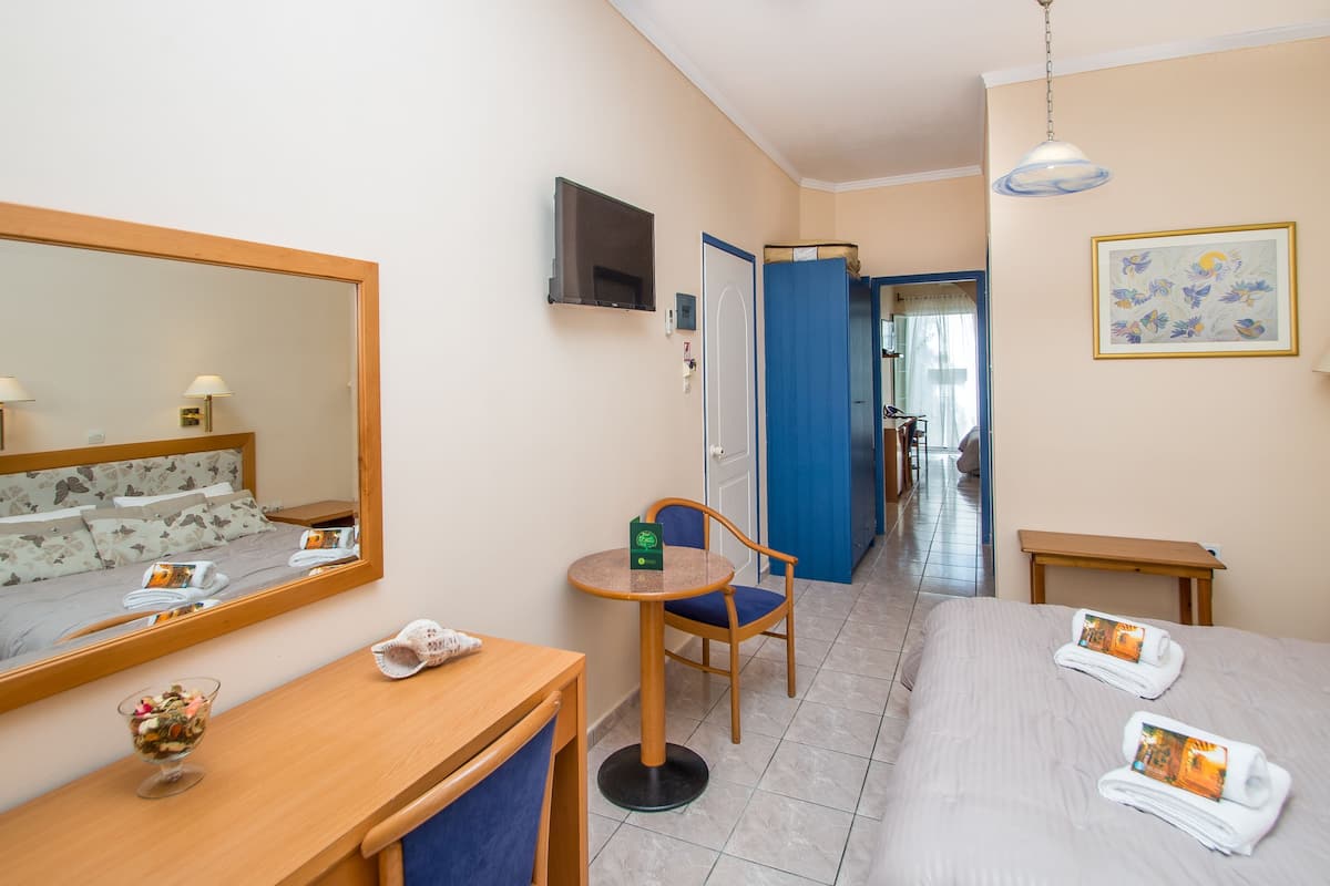 A hotel room with a bed, a desk, a chair, a small table, a mirror, a TV, and a door leading to another room.