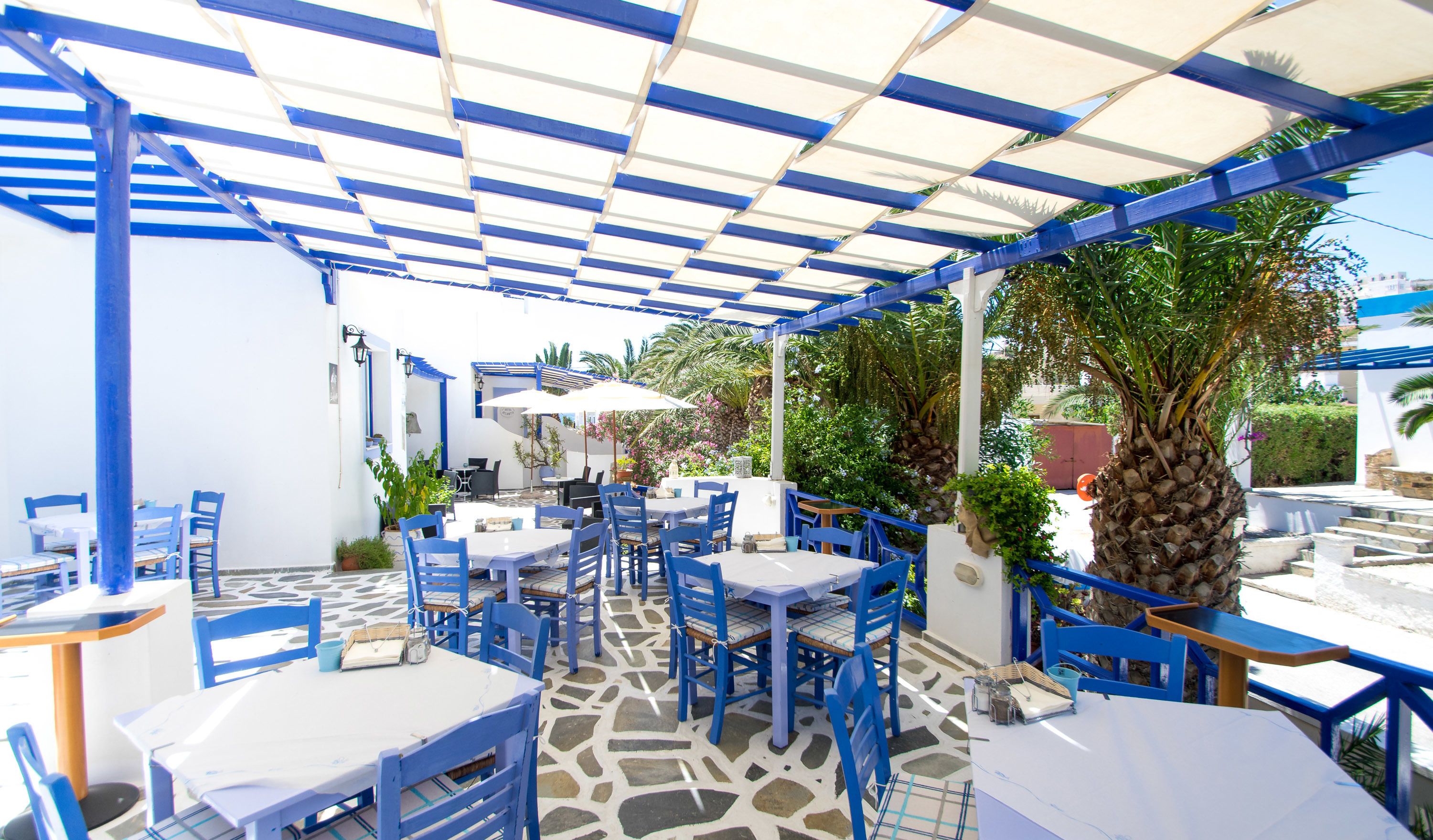 Un café en plein air avec des meubles bleus et des tables blanches, entouré de verdure et doté d’un sol à motifs.