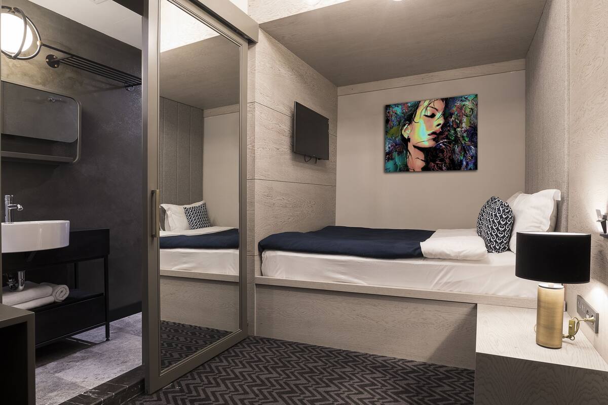 Habitación de hotel moderna con un espejo grande, una cama con ropa blanca, un cojín con estampado blanco y negro, un cuadro en la pared y una mesita de noche con una lámpara.