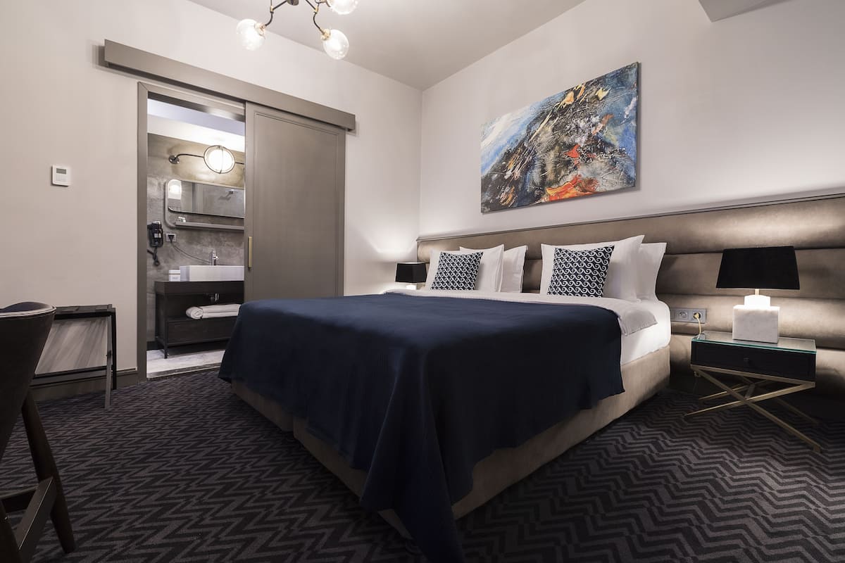 Una habitación de hotel moderna con una cama grande, mesitas de noche, un escritorio y un baño visible a través de una puerta abierta.