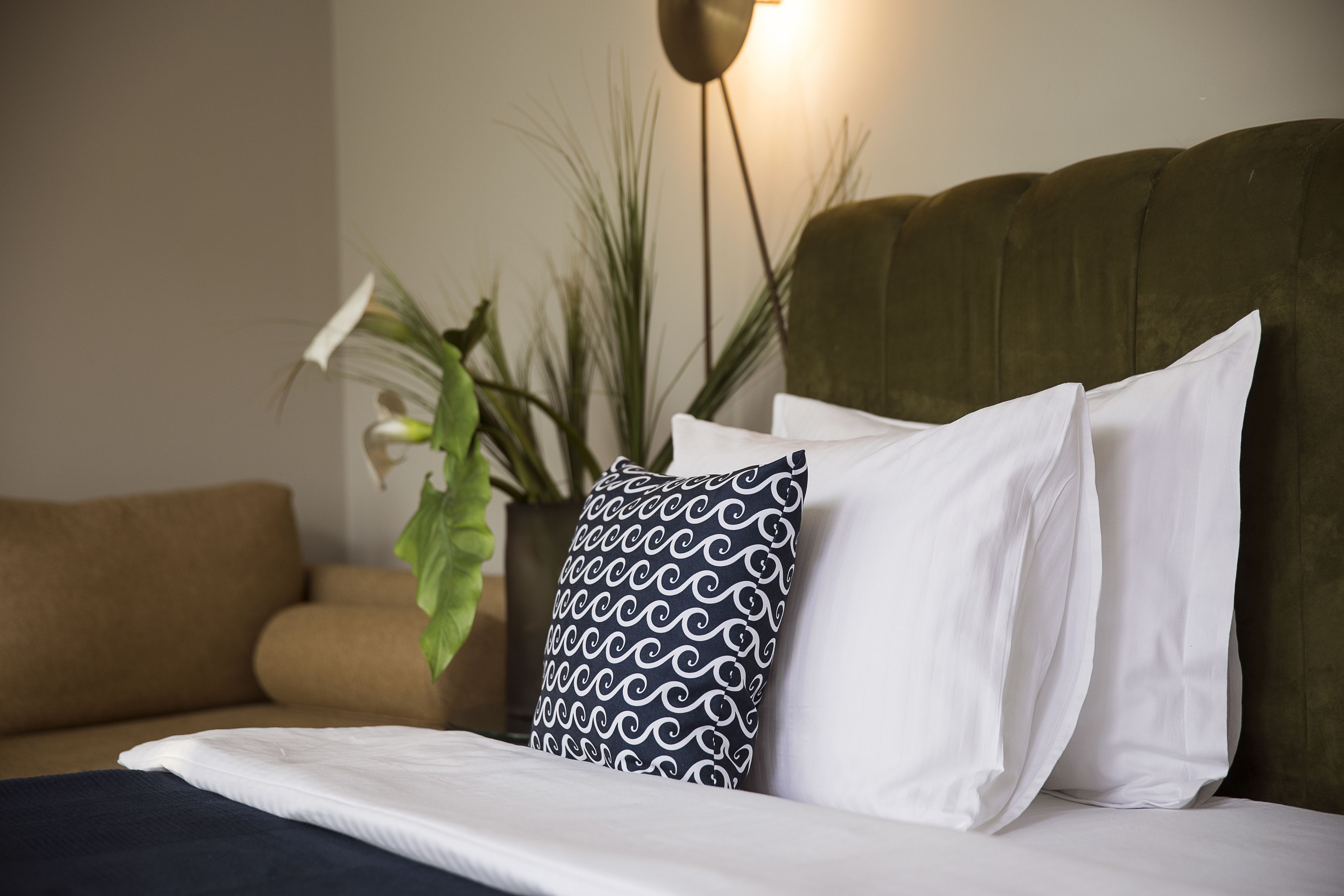 A neatly made bed with a patterned pillow, a green upholstered headboard, and a potted plant in the background.