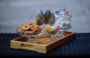 Una bandeja de madera con un tazón de vidrio para postres con galletas, otro tazón de vidrio con caramelos de colores y un recipiente de vidrio con caramelos blancos.