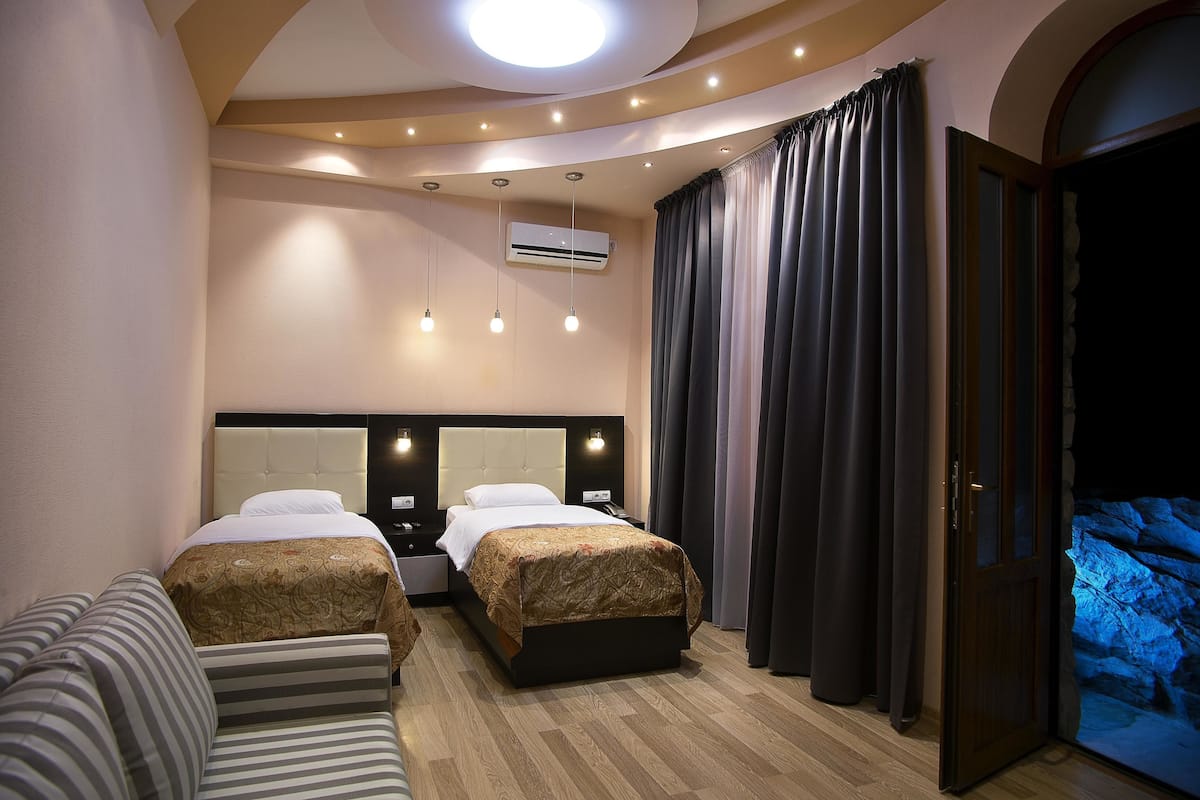 Ein Hotelzimmer mit zwei Betten, einem Sofa, einem großen Fenster mit Vorhängen und einer Wandverkleidung aus Stein.