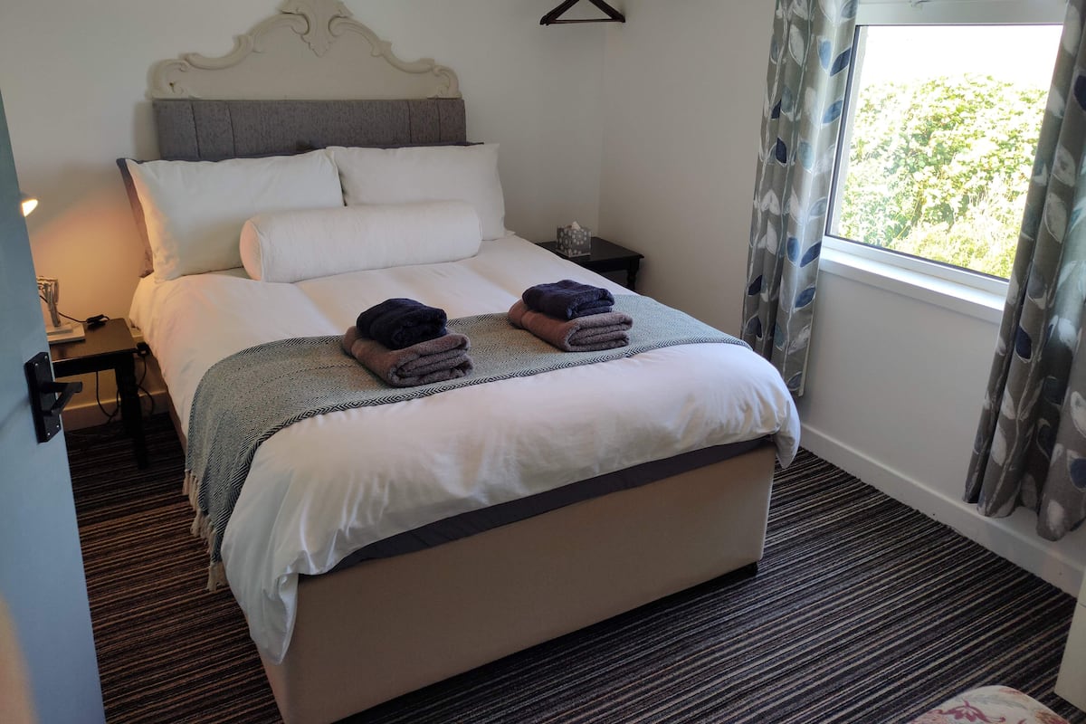 A neatly made bed with a patterned throw, a bedside table with a lamp, and a window with curtains.