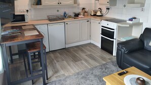 Een compacte keuken met witte kastjes, een magnetron, een vaatwasser en een fornuis. Er is een houten tafel met boeken en een afstandsbediening op een bijzettafeltje.