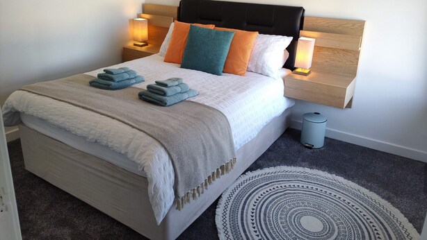 Een net opgemaakt bed met een hoofdbord, twee bedlampjes en een klein prullenbakje.
