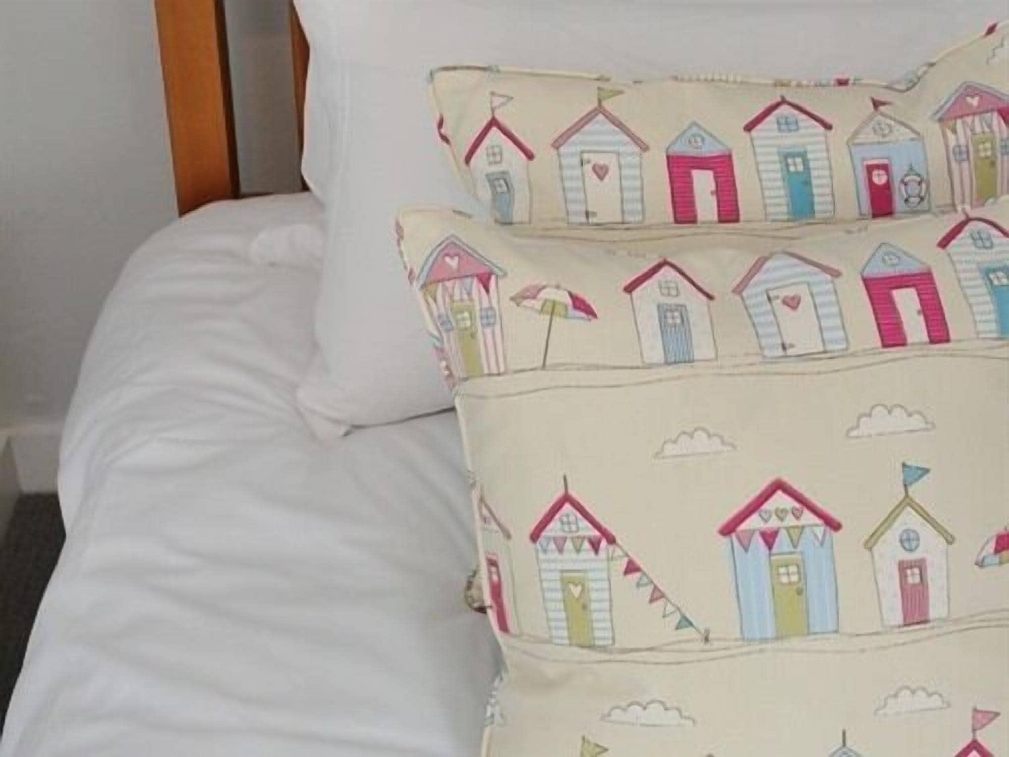 Un lit avec une literie blanche et des coussins décoratifs ornés d’un motif représentant un village au bord de la mer.