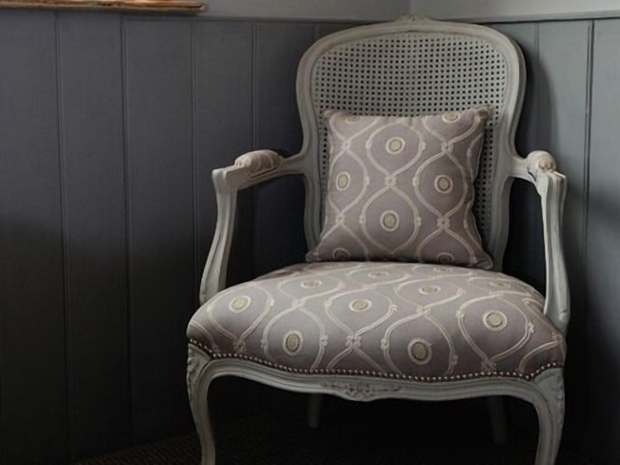 A vintage-style chair with a patterned cushion, upholstered in a light fabric, against a dark paneled wall.