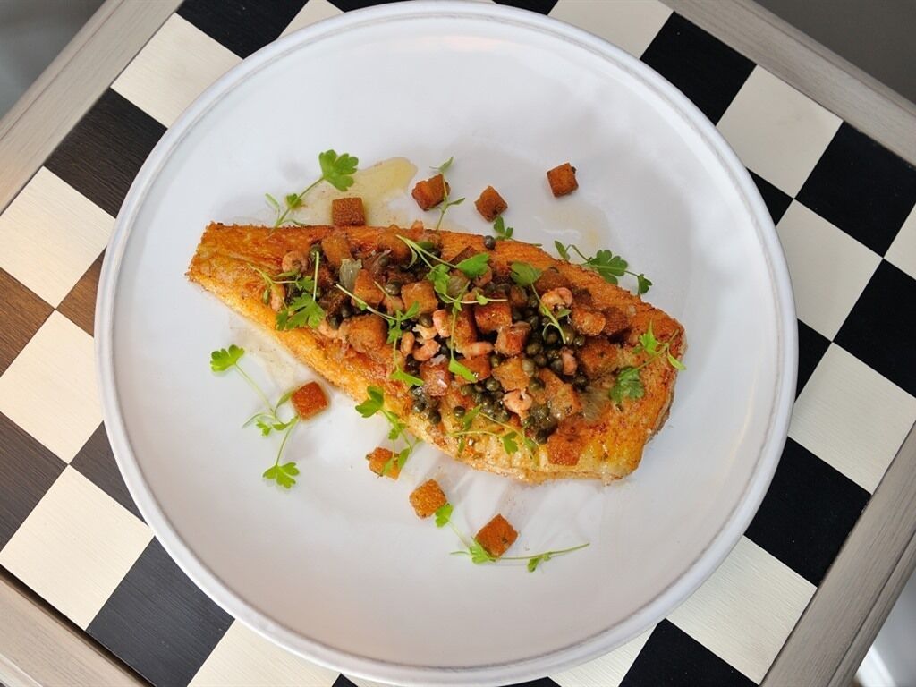 A white plate with a piece of food garnished with herbs and small cubes, placed on a checkered tablecloth.
