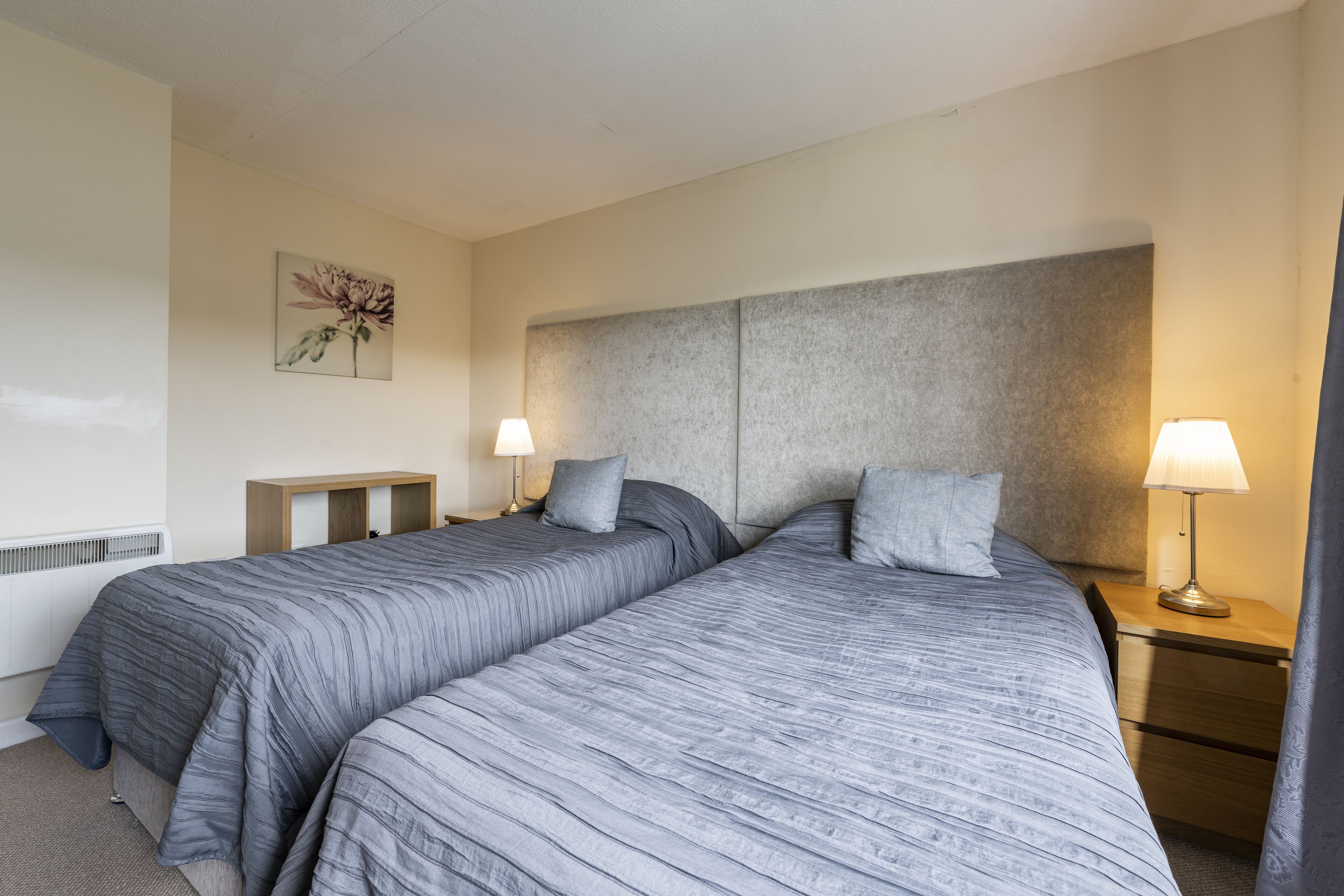 A hotel room with two beds, a bedside table with a lamp, a small wooden nightstand, and a wall-mounted artwork.
